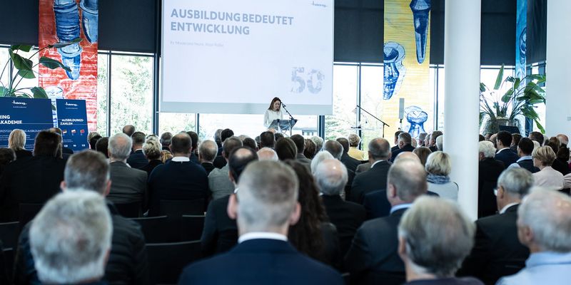 Publikum sitzt vor einer Bühne, junge braunhaarige Frau am Mikrofon, auf der Bühne ist eine Leinwand mit der Aufschrift "Ausbildung bedeutet Entwicklung"