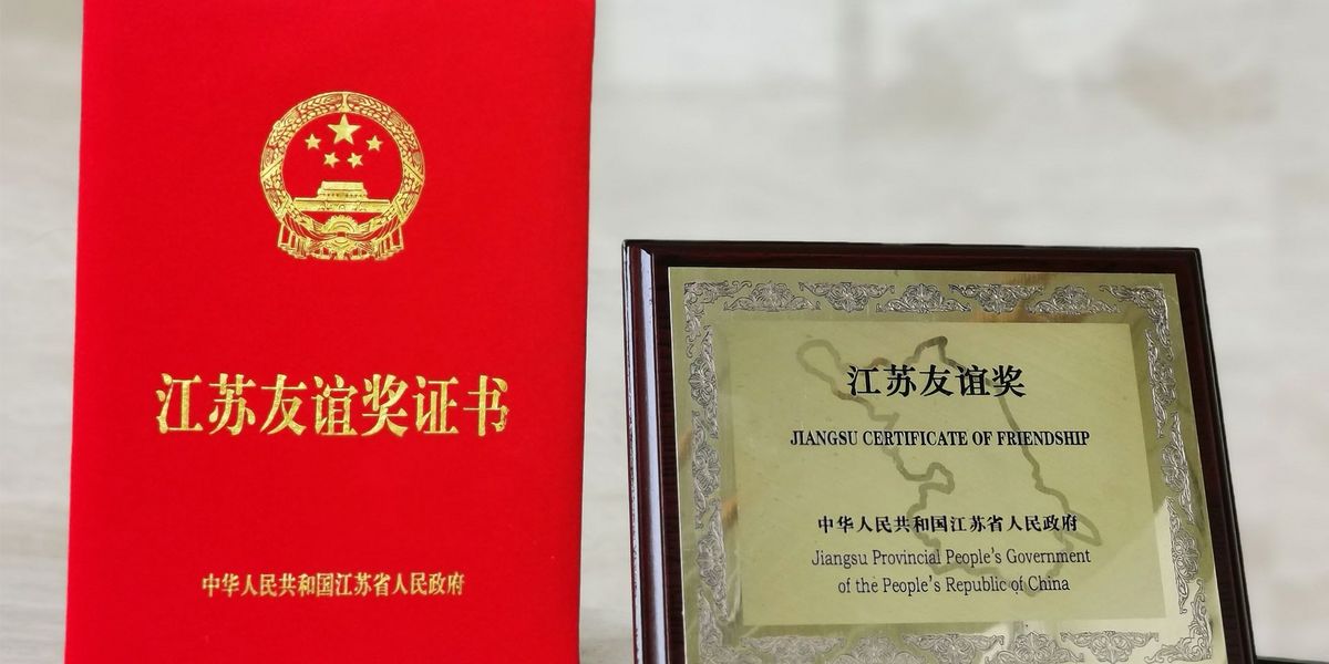 Freundschaftspreise, links rotes kleines Buch mit chinesischer goldener Aufschrift und rechts ein goldenes Bild mit holz Umrandung und schwarzer Schrift "Jiangsu Certificate of Friendship - Jiangsu Provincial People´s Government of the People´s Republic of China"